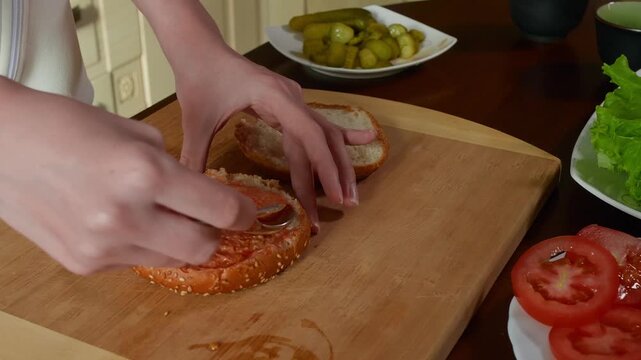 Hands spreading red pate or ketchup onto seeded bread slices on wooden cutting board with fresh vegetables, overhead camera perspective capturing food preparation detail.