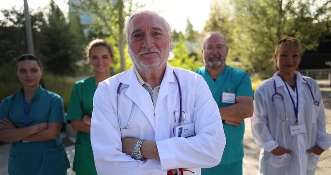 Senior male doctor with a white lab coat and stethoscope standing confidently with arms crossed in front of his diverse medical team, all looking at the camera with pride and expertise