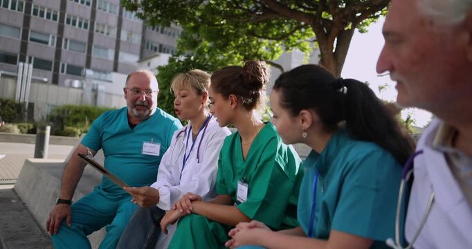 Professional team of doctors and nurses sitting together outside a hospital building, discussing a patient's case while reviewing medical records on a clipboard and having an informal meeting