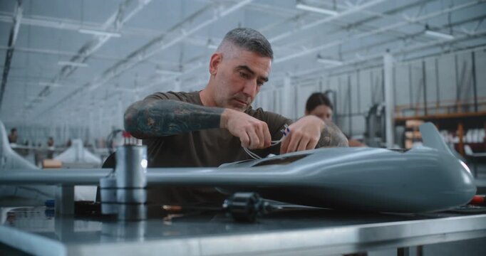 Male Technician Integrates Electronic Components, Cylindrical Module and Wiring inside Fixed Wing UAV During Detailed Drone Assembly Process in High Tech Modern Aerospace Manufacturing Facility.