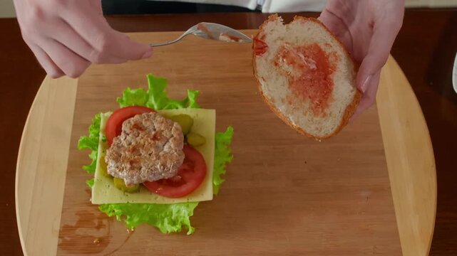 Close up of hands spreading ketchup pate onto toasted bread with knife while assembling sandwich with lettuce, tomato, cheese on wooden cutting board, kitchen preparation.