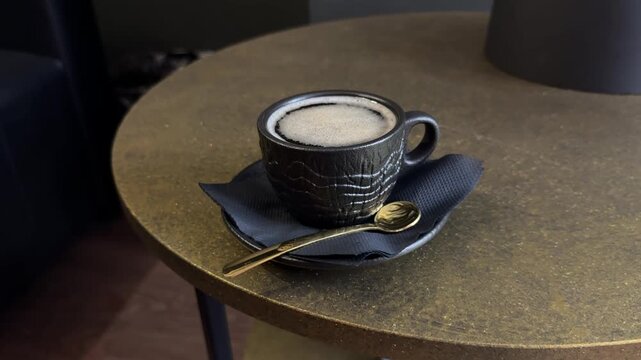 Horizontal video with gentle camera movement around a black ceramic cup of fresh foamy coffee on a textured round table, served with a golden teaspoon and dark napkin in a cozy minimalist interior