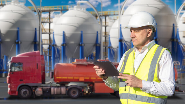 Fuel storage terminal. Engineer monitors tanker truck loading near industrial storage tanks for fuel distribution, logistics and terminal operations.