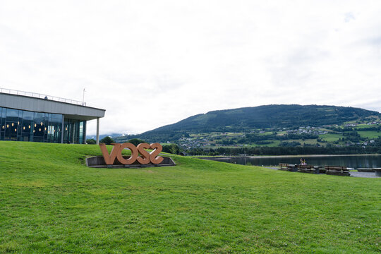August 17, 2026, Voss, Norway - Large 'VOSS' sign on a grassy hill with a modern building, lake, and forested mountains in the background