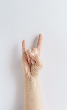 Vertical close-up shot of a human hand showing a rock and roll sign or devil horns gesture against a clean white backdrop, representing music, rock culture, and energy.