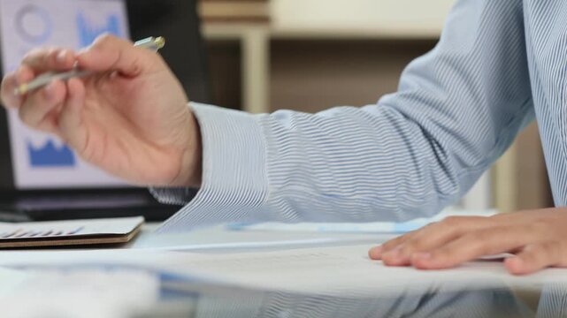Business person analyzing financial data, examining charts and graphs on documents and a laptop, and taking notes with a pen in a modern office environment