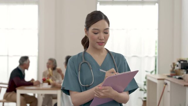 Female Asian caregiver reviews senior care area and records notes on clipboard while residents sit nearby in assisted living center, supporting routine inspection, safety and care management.