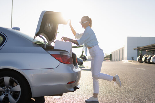 Woman loading white shopping bags into car trunk after successful black friday sales event, enjoying discounts and consumerism in a bright outdoor parking lot