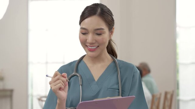 Female Asian caregiver looks around senior care area, pauses and writes notes on clipboard in assisted living center, supporting routine inspection, safety monitoring and organized care service.