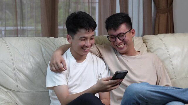 Two happy Asian young men sitting on a couch looking at a smartphone, laughing and sharing media together, friendship and technology concept