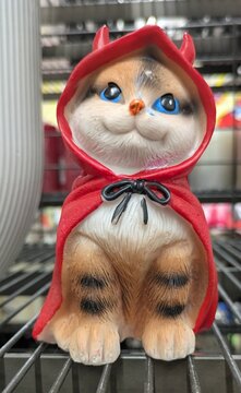 Tiempo de Halloween. Figura decorativa de un simp&aacute;tico gatito sentado, disfrazado de demonio con una capa con capucha con cuernos de color rojo, en estante de comercio