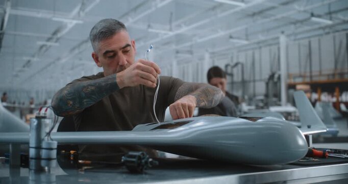 Skilled Male Specialist Installs Electronic Hardware, Cylindrical Unit and Connects Wiring Inside Fixed Wing Drone, Demonstrating Precision UAV Assembly in Modern Aerospace Production Facility.