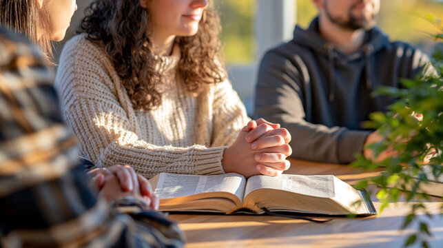 Over the shoulder view focusing on an open Bible surrounded by praying hands from a small group, faces cropped beyond the frame, bright clean room with soft window illumination, natural wood surface