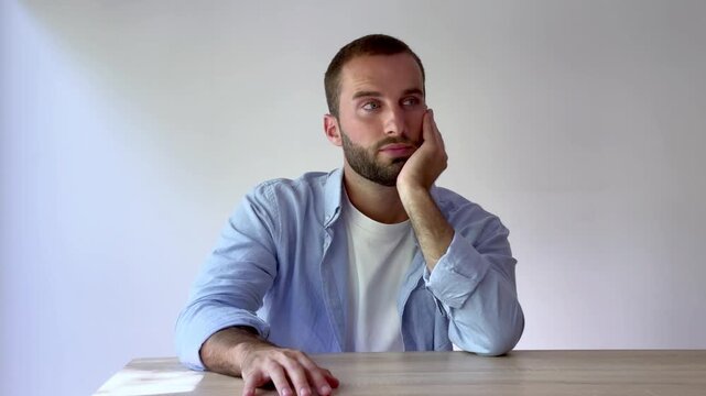 Bored and pensive young man sitting at a table, leaning on his hand and looking around with an impatient expression, feeling loneliness and frustration isolated on a white background