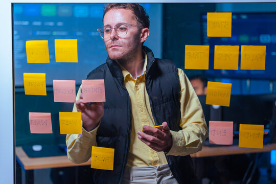 Focused professional wearing glasses placing sticky note on glass wall during project planning and idea strategy meeting in modern dark office