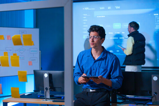 Focused software developer holding a tablet in a modern tech office with workflow planning boards and screens in the background
