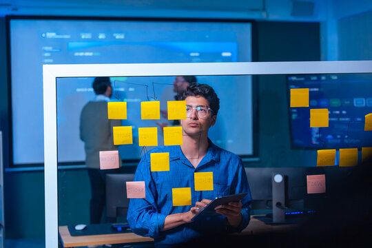 Male developer looking through glass board while holding tablet to plan software flowchart with sticky notes in tech company