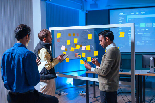 Diverse developers team analyzing project strategy and writing notes during effective planning meeting near glass board in modern tech office