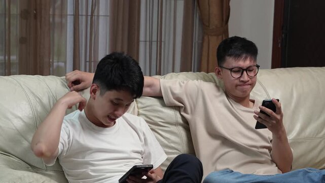 Two Asian young men sitting on a couch using smartphones separately, looking bored and disconnected, digital addiction and apathy concept