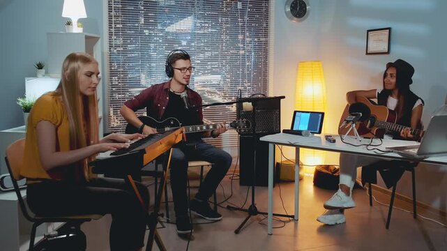 Four young adults playing acoustic guitars and keyboard together in a home recording studio with musical equipment and cityscape view through window blinds.