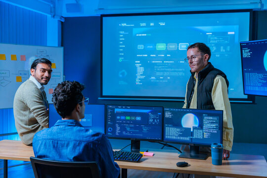 Diverse software development team analyzing system performance and coding data on multiple screens in modern technology control room