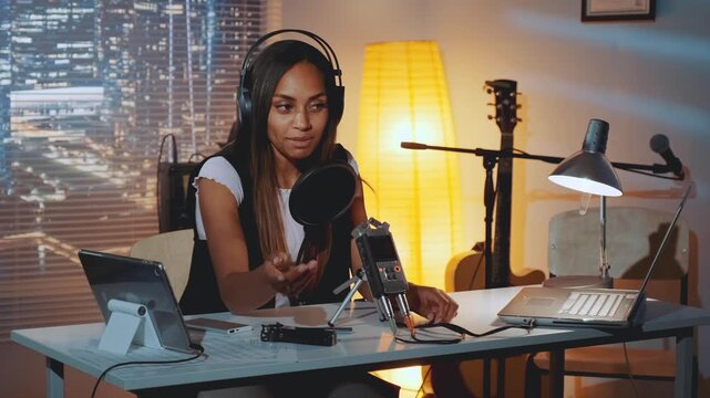 A female journalist wearing headphones is broadcasting an online interview from a well-equipped home recording studio at nighttime with city lights visible outside.