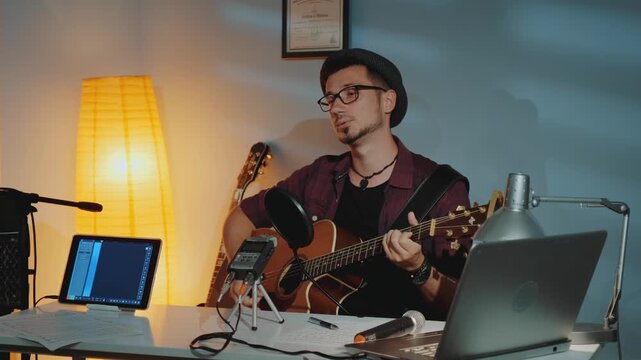 A young man wearing glasses and a fedora hat plays an acoustic guitar in a music studio surrounded by recording equipment and instruments indoors.