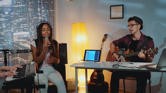 Three musicians collaborate in a home studio at nighttime, with one singing into a microphone, another playing acoustic guitar, and a third on keyboard synthesizer.
