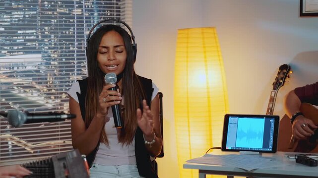 A black woman with long dark hair sings passionately into a microphone wearing headphones in a home studio with musical instruments and city lights at night.