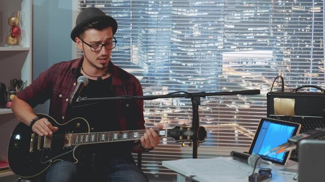 A young man wearing a fedora and glasses plays acoustic guitar and sings into a microphone in a nighttime home recording studio with city lights visible outside.