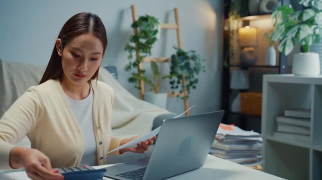Young Asian woman calculates expenses and checks bills with laptop in living room at home. Personal finance budget planning, Tax return invoice paperwork, Household banking savings analysis concept.