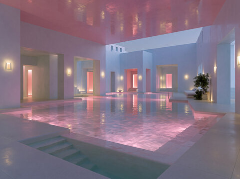 Dreamy pink indoor pool space with textured plaster walls, multiple arches, glowing wall lamps and shimmering pink lit pool water.