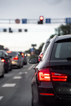 Passenger cars queued at a red traffic signal with illuminated rear lamps and overhead lane signs, representing intersection control and delays in everyday road transport