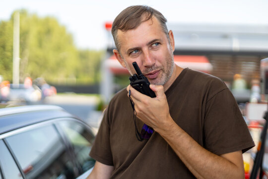 Man Using Handheld Radio Beside a Car. Middle-aged man holding a handheld two-way radio near his mouth while standing beside a car outdoors.
