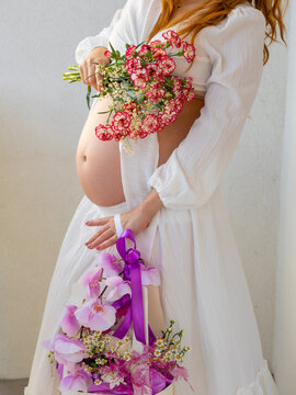 Pregnant woman in white dress holding colorful flowers during romantic pregnancy photoshoot. Romantic maternity photograph featuring a pregnant woman in a white dress holding colorful flowers near her