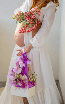 Pregnant woman in white dress holding flowers during elegant maternity photo session. Elegant maternity portrait of a pregnant woman wearing a flowing white dress and holding a colorful bouquet 