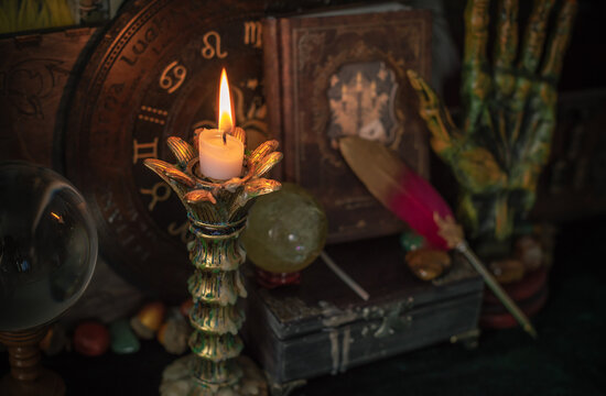 Burning candle in vintage candlestick on mystical altar with crystal ball, zodiac wheel, old spell books and dip pen 