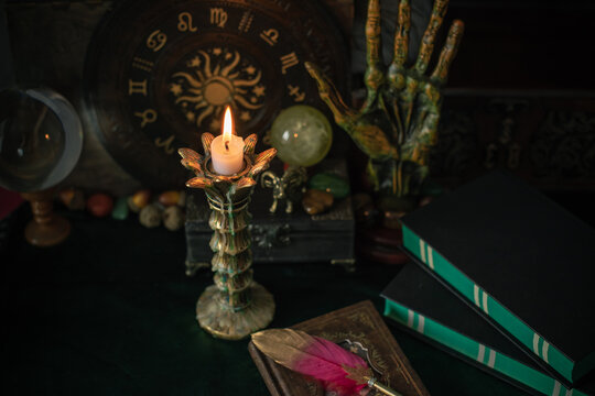 Burning candle in vintage candlestick on mystical altar with crystal ball, zodiac wheel, old spell books and dip pen 