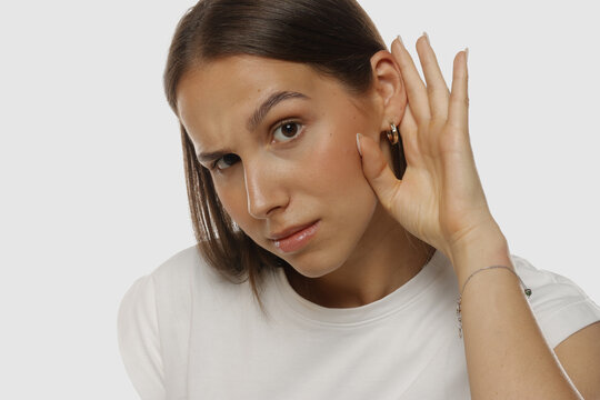 Portrait of a young woman listening carefully with hand near her ear, natural beauty, no makeup, on white background.