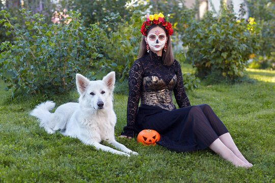 Halloween portrait of woman sitting on grass with white dog and Halloween pumpkin in garden.