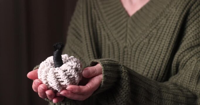 Person holding knitted pumpkin with black stem in hands, olive-green sweater, studio setup, autumn decor, handmade styling
