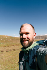 Man with beard and backpack smiles at camera in open field. Blue sky above grassy hills. Outdoor adventure, hiking, travel, or wellness theme. Could use in tourism, fitness, or nature content © Pauli