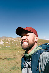 Man with beard smiles in open mountain meadow. He wears red cap and hiking backpack. Sunny day highlights his joyful expression. Ideal for travel, adventure, or outdoor lifestyle themes © Pauli