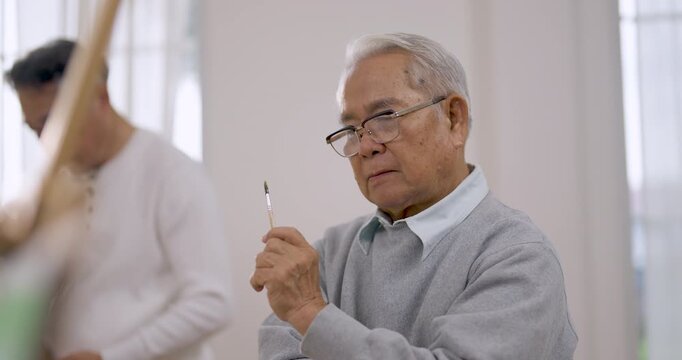 Senior Asian man studies brush before painting at easel during art activity in assisted living community, supporting creative expression, calm relaxation, emotional wellbeing and active aging.