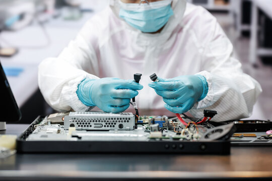 Technician in protective clothing and gloves repairs an open laptop, holding small cable connectors over exposed computer parts in a clean electronics workshop.