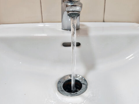 Water flowing from chrome faucet into white ceramic bathroom washbasin. Sink drain hole with active water stream