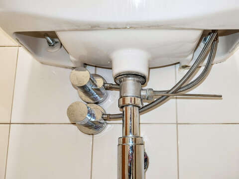 Plumbing connection under white bathroom sink. Chrome faucet water supply pipes and drain hardware. Maintenance of washbasin assembly for residential or public restroom facility