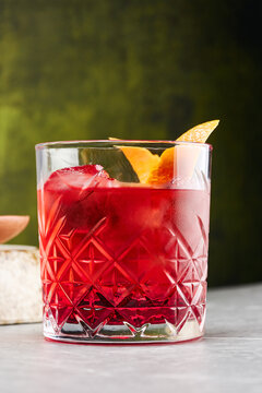 Deep red negroni cocktail with orange twist and ice against a moody green wall, drink close up