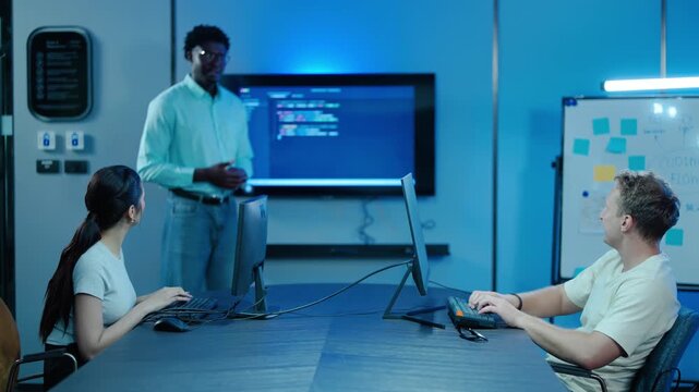 Diverse software developer team collaborating in a modern tech office. Programmers are actively coding and discussing a complex project on a large screen with blue lighting for IT teamwork concepts.
