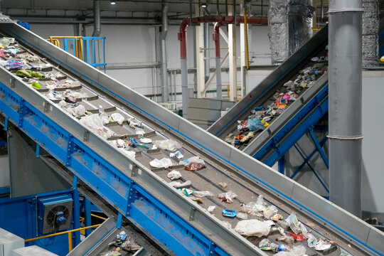 Blue machinery carries piles of discarded plastic and trash along rising metal belts within a large warehouse setting dedicated to environmental sorting